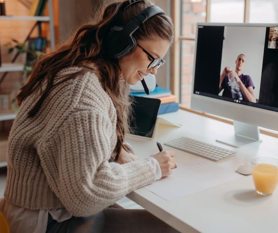 A woman on a one-on-one video call smiling and taking handwritten notes