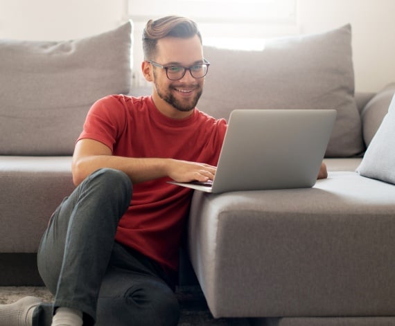 A man casually sitting next to his couch at home, working on his laptop and smiling