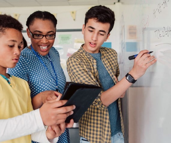 Three students working together as one writes something on a whiteboard and another points at something on a tablet