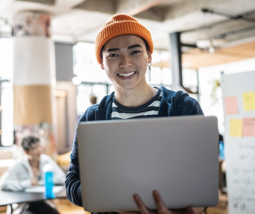 A young person wearing a beanie and holding a laptop while smiling at the camera