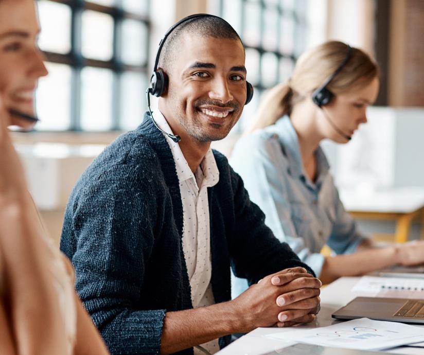 A photo of multiple people working at a desk wearing headsets, and the man in the middle is looking up at the camera and smiling