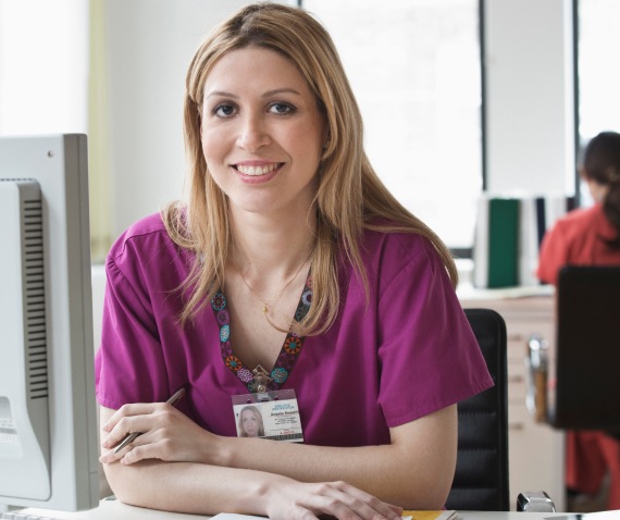 A women in scrubs sitting at a desk, leaning forward and smiling