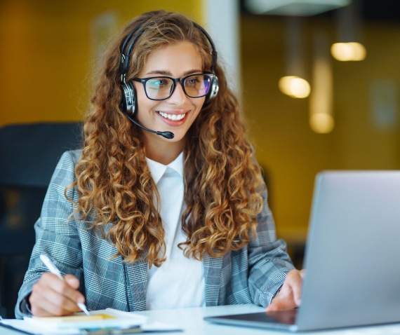 A woman wearing a headset smiling and looking at her computer while holding a pen for note-taking